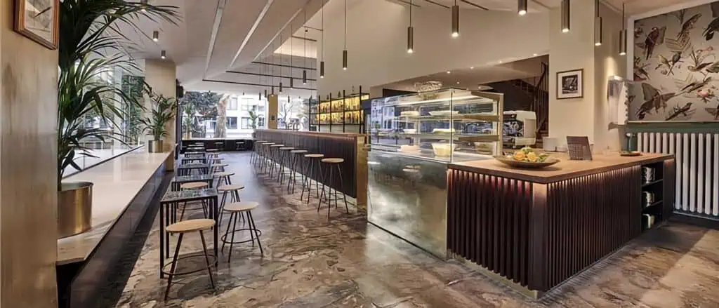 Innenansicht des Clubs „3sein“, das eine moderne und stilvolle Atmosphäre mit Holz- und Metallakzenten zeigt. Ein langer, eleganter Barbereich mit Hockern lädt zum Sitzen ein, während im Hintergrund ein Kühlregal mit frischen Snacks und Getränken zu sehen ist. Die Wände sind mit einer kreativen Tapete mit Vogelmotiven verziert, und warmes Licht aus Pendelleuchten beleuchtet den Raum.