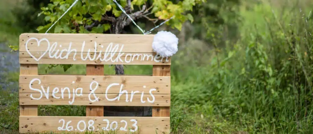 Ein liebevoll gestaltetes Holzschild mit der Aufschrift „Herzlich Willkommen Svenja & Chris – 26.08.2023“ empfängt die Gäste im Grünen.