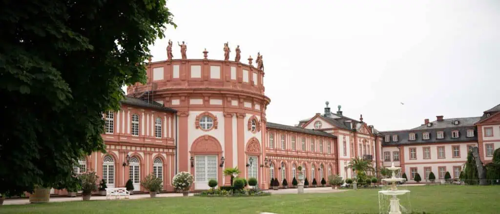 Außenansicht des Schlosses Biebrich in Wiesbaden, dem Veranstaltungsort der Abschlussfeier des Informatikstudiengangs der HSRM 2023. Die barocke Schlossanlage mit ihrer markanten roten und weißen Fassade erstreckt sich um einen gepflegten Garten mit einem Springbrunnen. Links im Bild rahmt ein großer Baum das historische Gebäude ein.